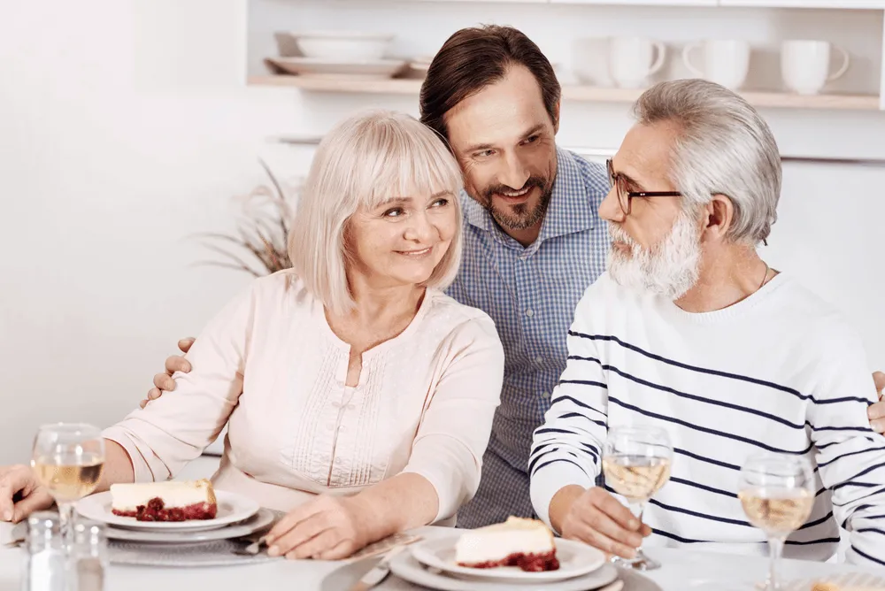 Elderly couple having cheesecake and wine with younger man smiling between them.
