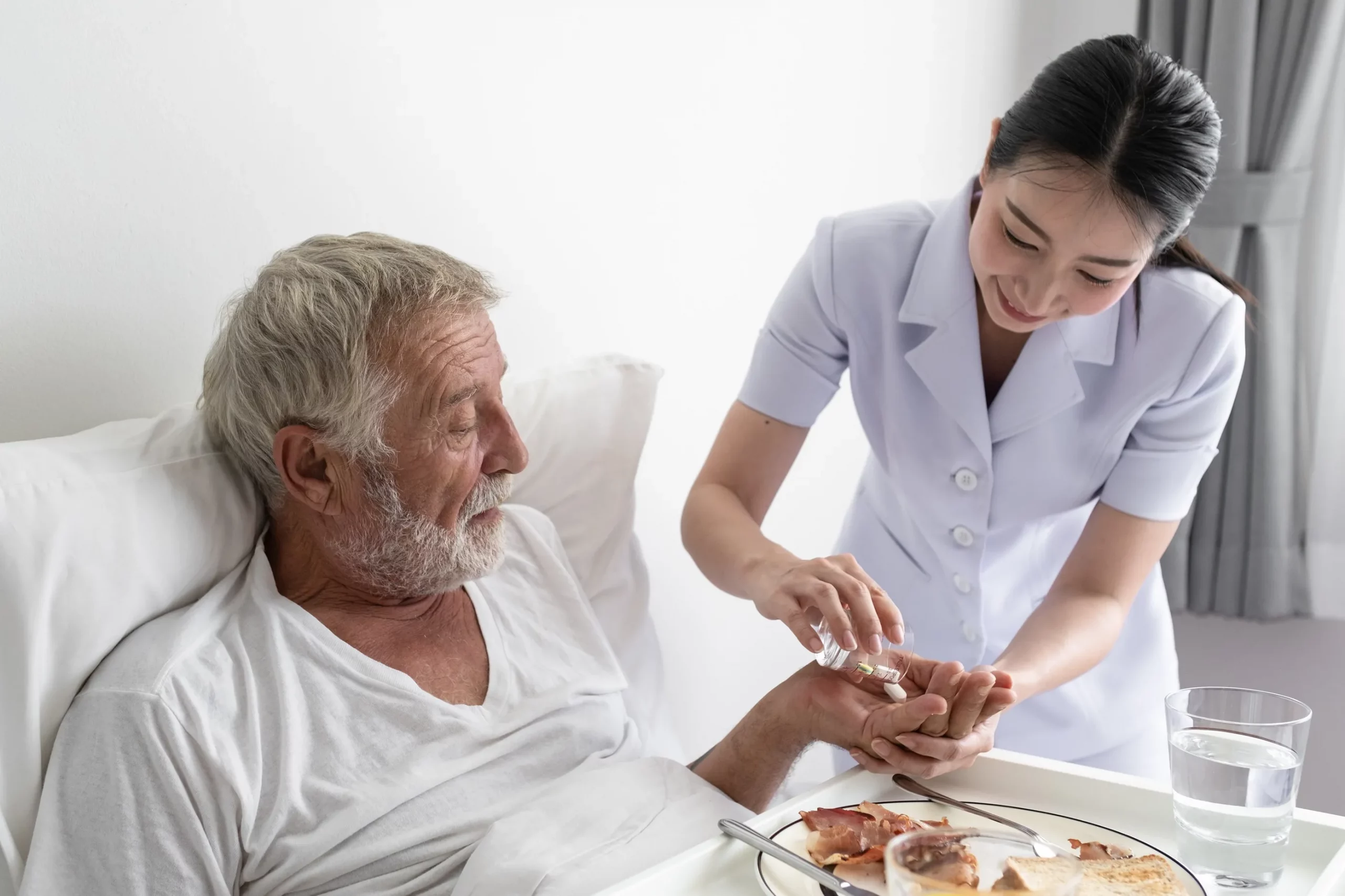 A home health aide giving medicine to an elderly man in bed.