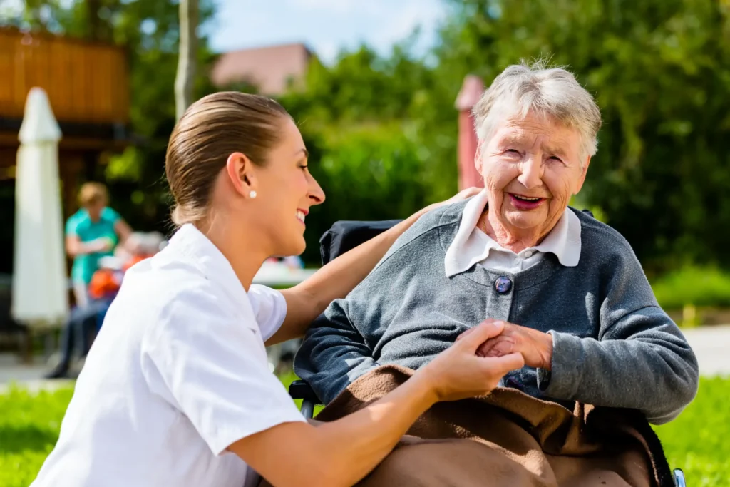 An elderly woman smiling in a wheelchair outside holding hands with her home health aide.