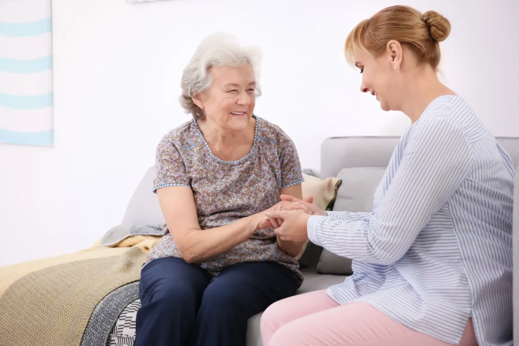 A young woman holding the hand of an older woman while sitting on the couch, both women smiling.