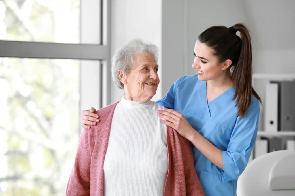 A home health aide smiling at her patient with her arms on her patient's shoulders.