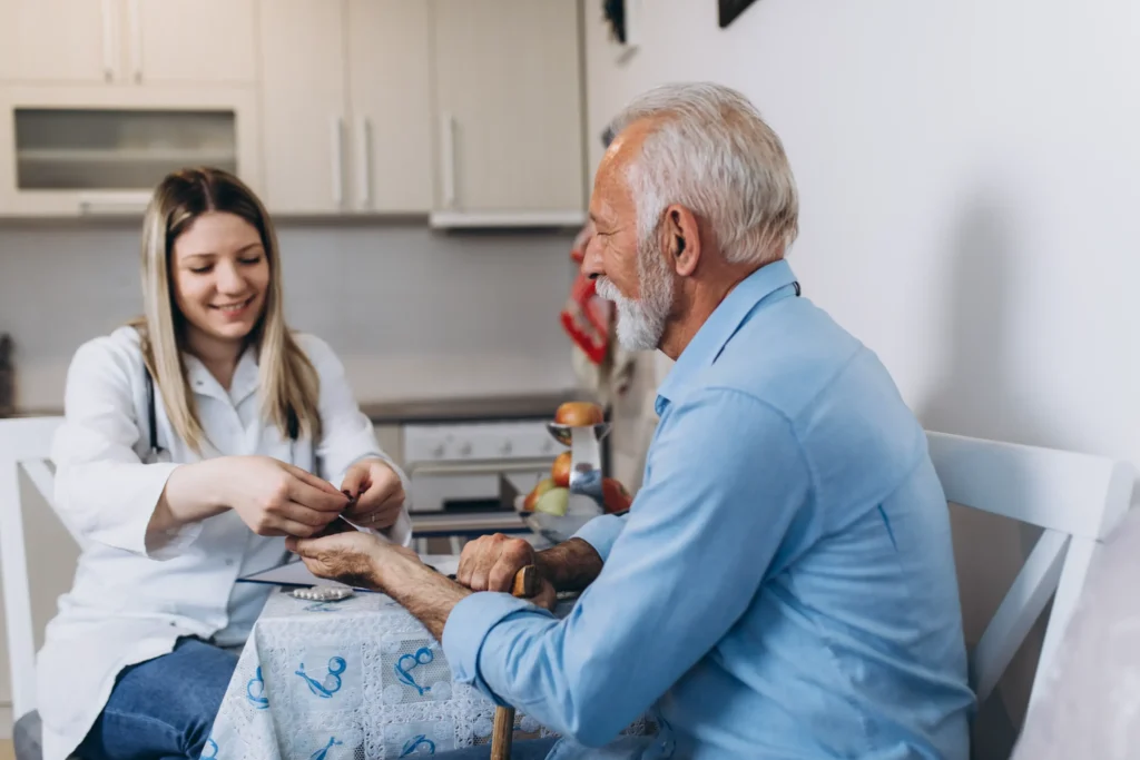 A home health aide giving medications to an elderly man in his kitchen.