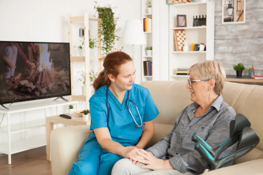 A home health aide talking to a woman on the couch with her arm support crutches next to them.