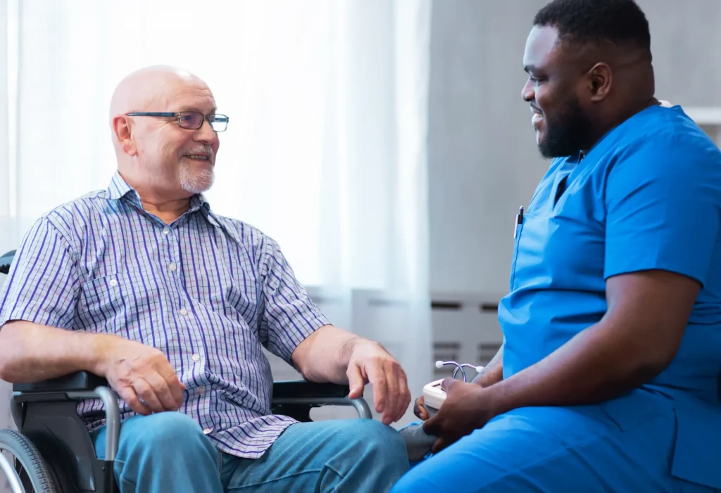 A man in the wheelchair smiling at his home health aide holding medical instruments.