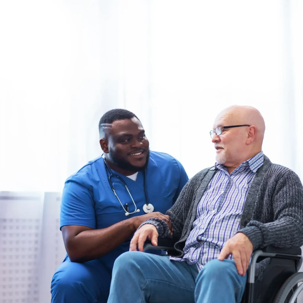 A smiling home health aide speaking to his patient who is sitting in his wheelchair.