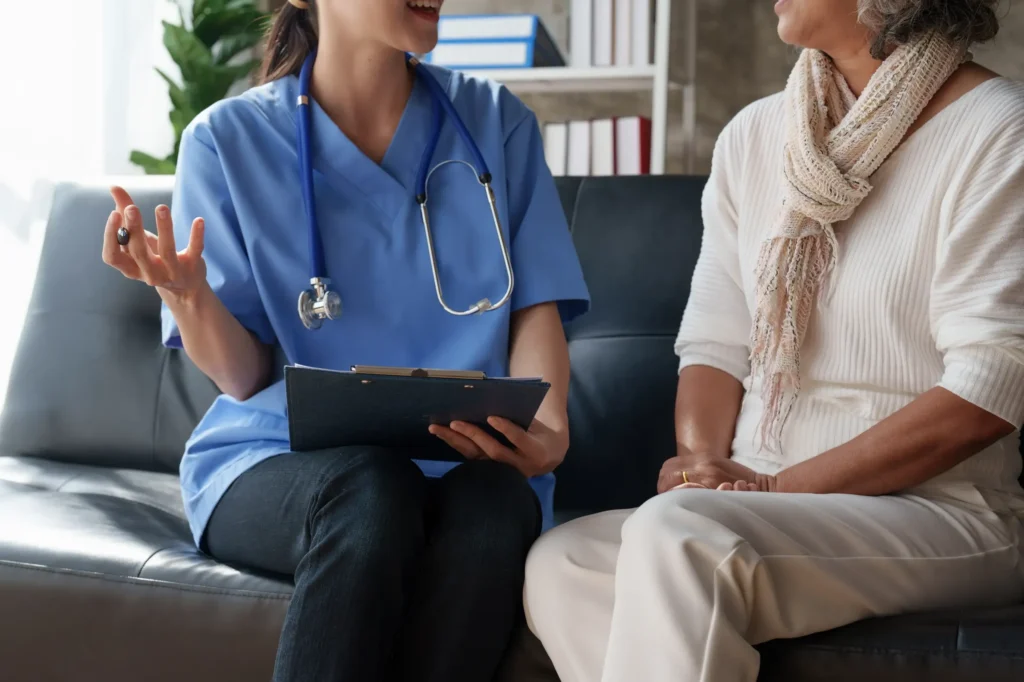 A home health aide is sitting on the couch with her patient speaking while holding a pen and clipboard.