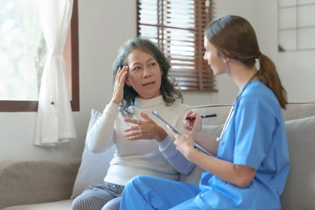 An elderly woman pointing to her head and talking to a health provider who is taking notes on a clipboard.