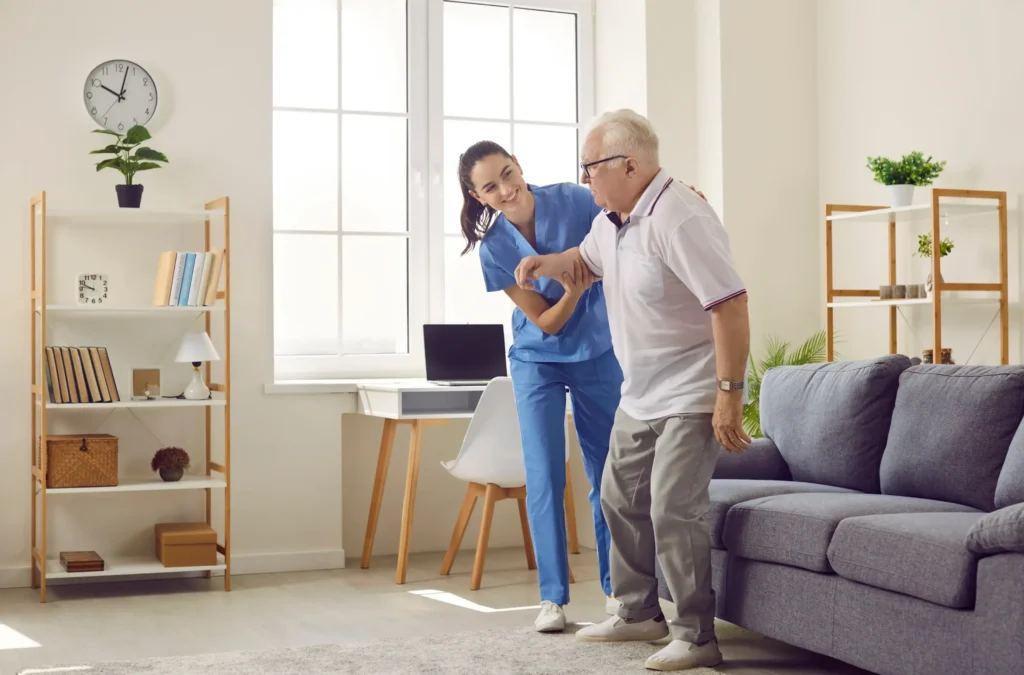 A home health aide helping up an elderly man from the couch.