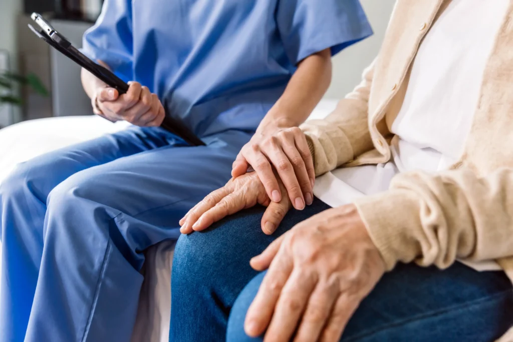 A home healthe aid holding an iPad and resting her hand on her elderly patient's hand.